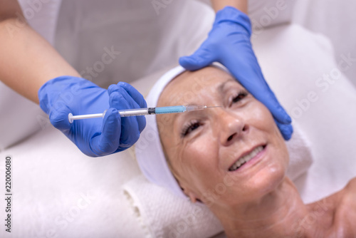 Syringe with hyaluronic acid and senior woman during skin treatment in the background