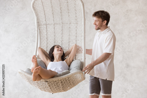 Smiling millennial boyfriend swing excited girlfriend in hanging lounge chair at home, happy woman relaxing in cozy suspended sofa while her lover dandle it, couple laughing having fun together