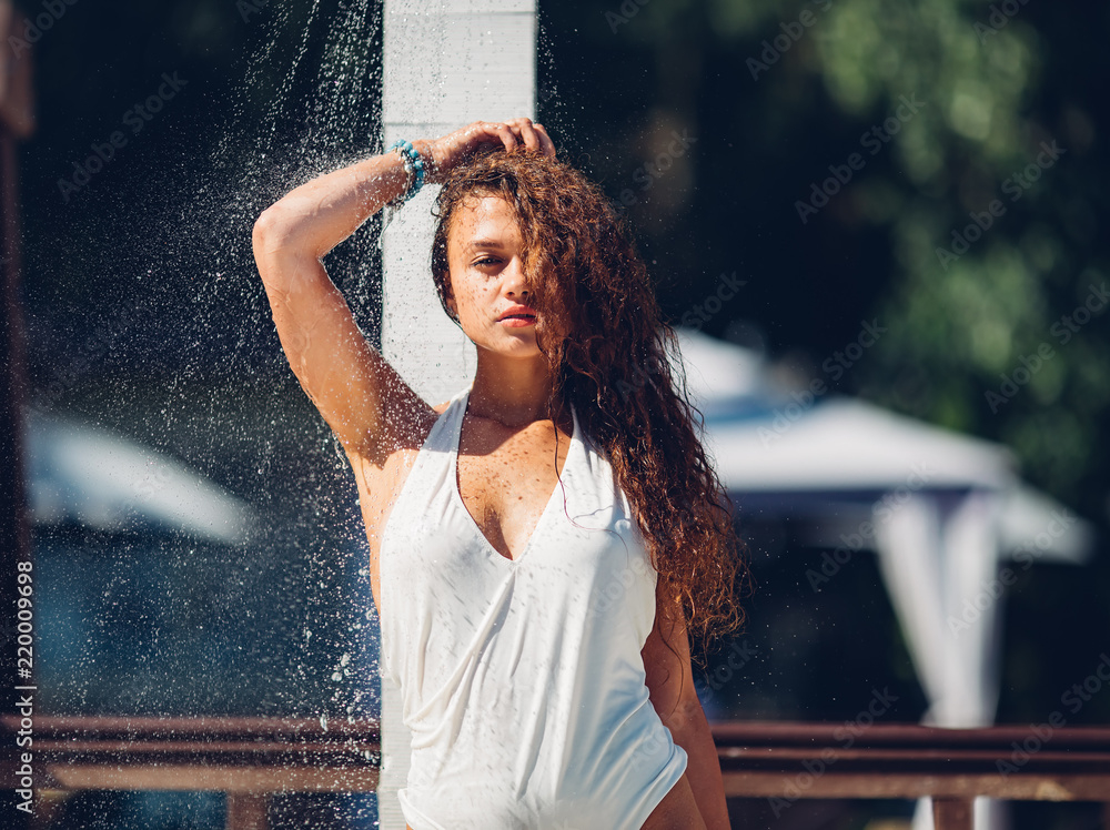 Heat and refreshment concept. Water drops falling on attractive woman. Young beautiful brunette girl taking shower on beach Sexy lady in bikini enjoys shower