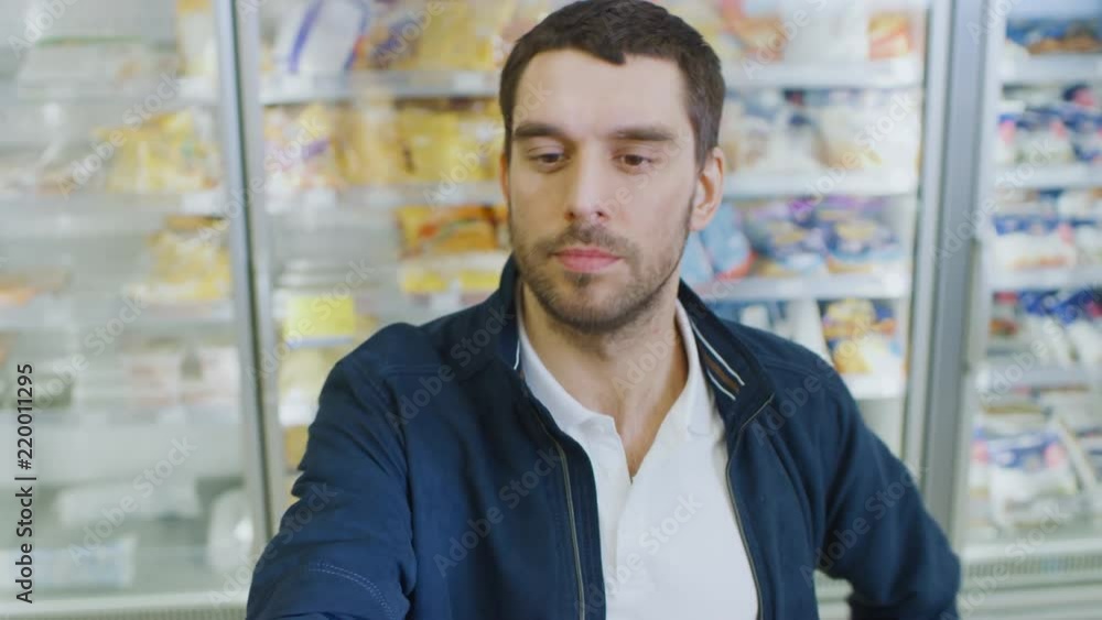 Portrait Shot of the Handsome Man Choosing Tin Can from the Canned Goods Section and Places it In His Shopping Cart. In the Background Frozen Goods Section of the Store. Portrait Shot of the Handsome Man Choosing Tin Can from the Canned Goods Section and Places it In His Shopping Cart. In the Background Frozen Goods Section of the Store.