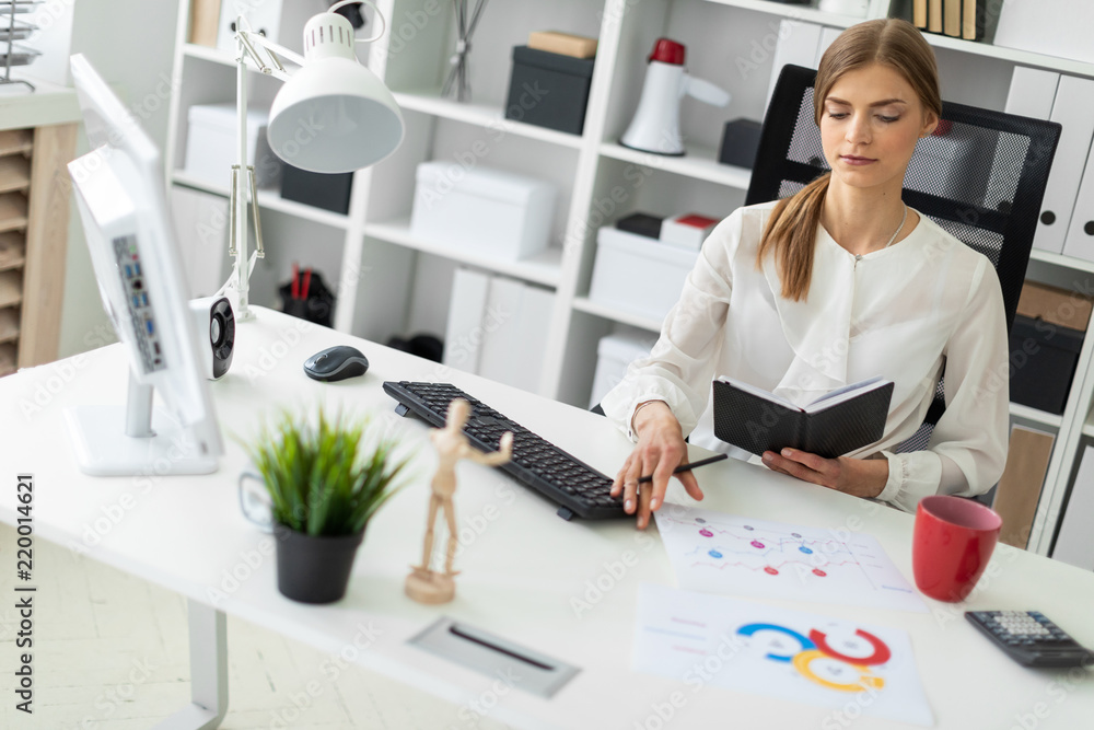 A young girl is sitting at a table in the office, holding a notebook and a pencil in her hand. Before the girl there are sheets with diagrams.