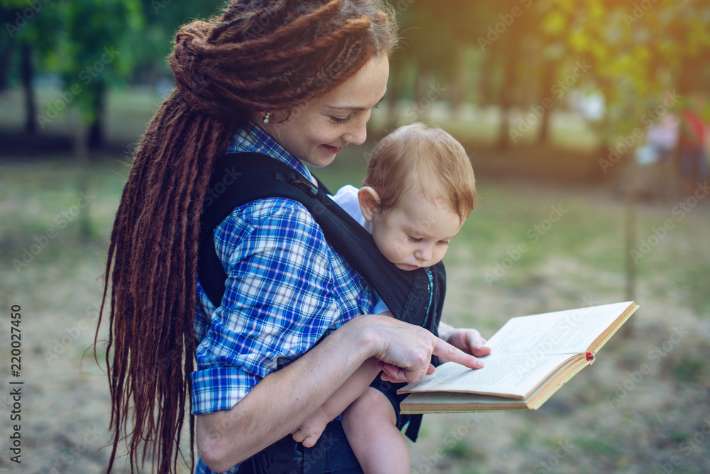 Mom with a baby in a sling reading a book in the Park. Concept of children's education in family life Mom with a baby in a sling reading a book in the Park. Concept of children's education in family life