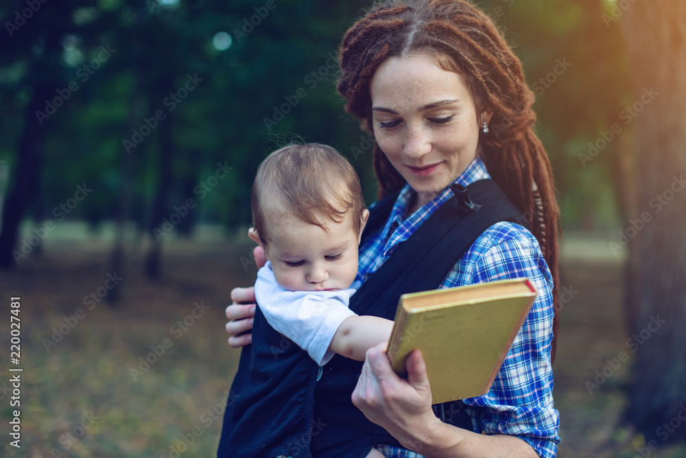 Mom with a baby in a sling reading a book in the Park. Concept of children's education in family life Mom with a baby in a sling reading a book in the Park. Concept of children's education in family life