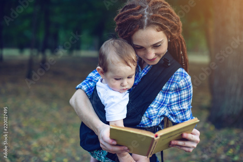 Happy mom with a baby in a sling reading a book in the Park. Children's education in family life