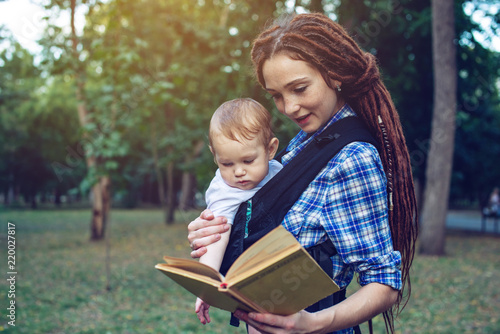 Beautiful mom with a baby in ergo backpack reading a fairy tale. Concept of children's education in family life © Artem