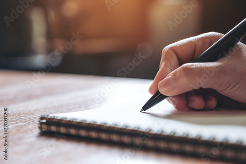 Closeup image of a hand writing down on a white blank notebook on wooden table