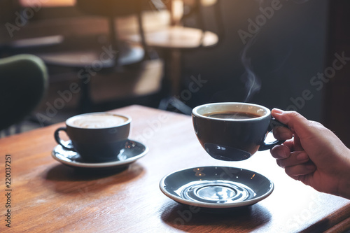 Closeup image of a hand holding a blue cup of hot coffee with smoke on wooden table in cafe