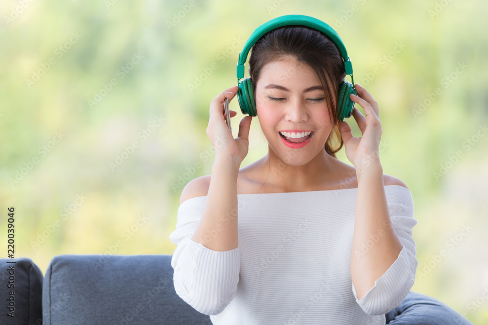 Close up shot portrait of young beautiful Asian woman sitting on sofa and listening to music with headphone in relaxing manner and hold smartphone in hand. Green nature blur in background Close up shot portrait of young beautiful Asian woman sitting on sofa and listening to music with headphone in relaxing manner and hold smartphone in hand. Green nature blur in background