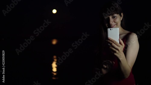 A young woman makes a video call on the background of a red plenilune, a rare astronomical phenomenon. The girl smiles and waves her hand. The orange moon is reflected in the river. photo