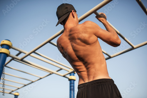 Rear view of young fitness muscular man doing pull ups exercises on horizontal bar outdoors. Handsome athletic shirtless male training hard at afternoon outside, sportsman working out outside.