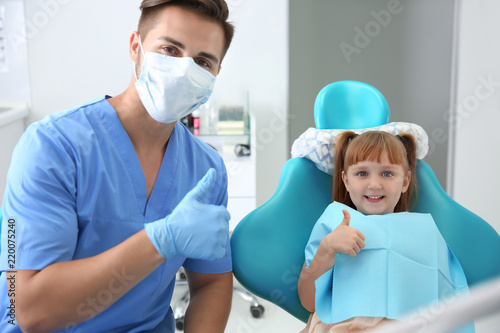 Cute little girl and dentist showing thumb-up gesture in clinic