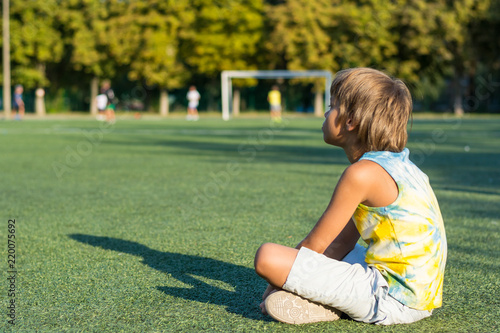 A boy is sitting on the grass at the edge of a school stadium and watching football
