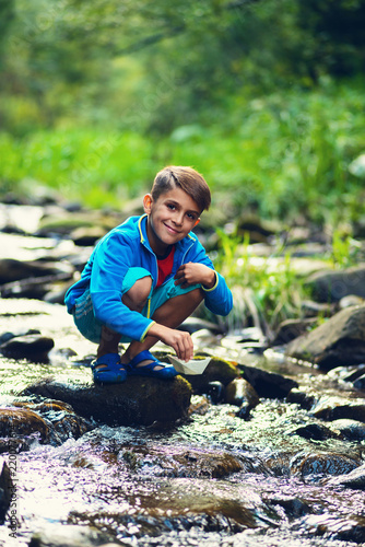 Obraz na płótnie Small boy sits on the rocks next to the mountain river and smiling w salonie