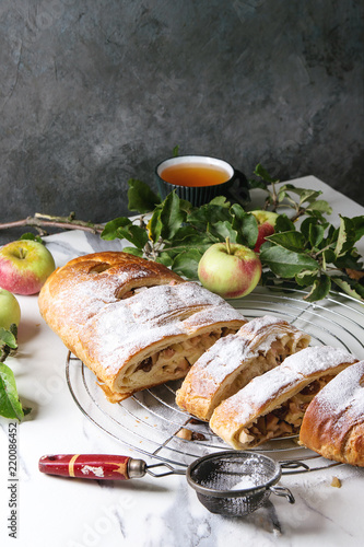 Homemade sliced puff pastry apple strudel pie on cooling rack served with ripe fresh apples, branches, cieve and sugar powder over white marble texture background.