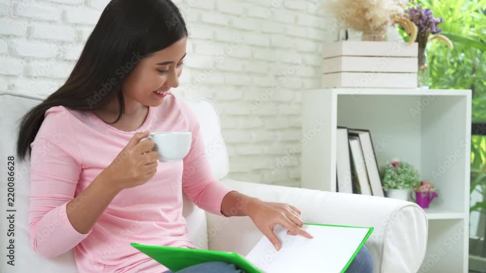 Young Asian attractive woman reading interesting book and drinking hot tea or coffee from a white cup on the couch in the cozy living room at home. Casual lifestyle at home. Young Asian attractive woman reading interesting book and drinking hot tea or coffee from a white cup on the couch in the cozy living room at home. Casual lifestyle at home.