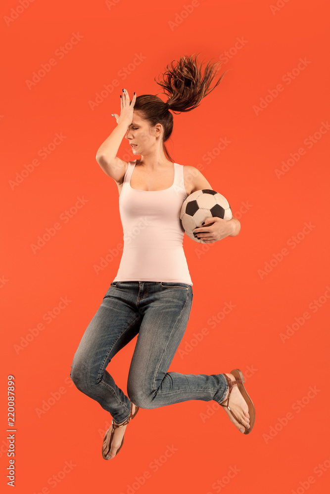 Fototapeta premium Forward to the victory.The young woman as soccer football player jumping and kicking the ball at studio on a red background. Football fan and world championship concept. Human emotions concepts