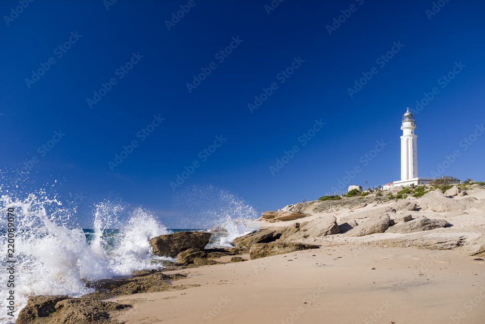 lighthouse on trafalgar beach with waves breaking on rocks in a sunny day with blue sky