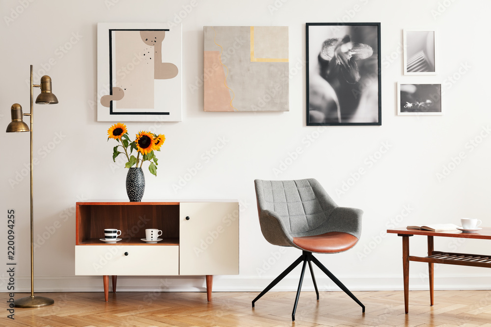 Fototapeta premium Grey armchair next to white cupboard with sunflowers in apartment interior with posters. Real photo