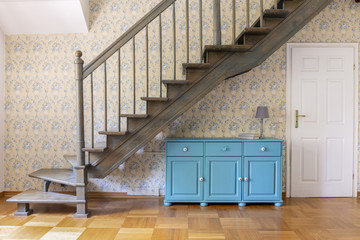 A light blue three door cabinet standing under gray staircase against a wall with flower wallpaper in a hall interior. Real photo.