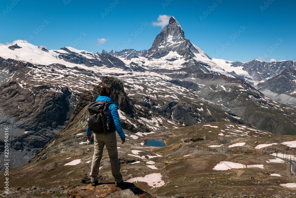 Female traveler with backpack hiking mountain trail and admiring views of Majestic Matterhorn mountain in Valais (Pennine) Alps, Switzerland.
