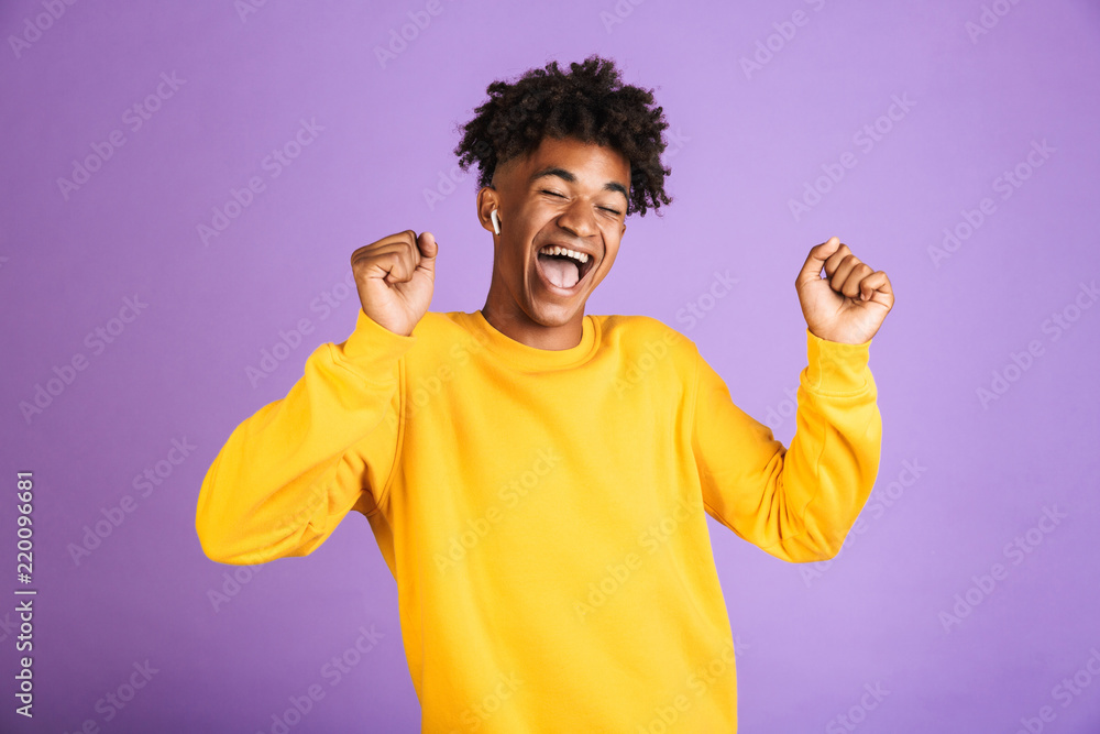 Portrait of happy african man having stylish afro hairdo dancing and singing, while listening to music via bluetooth earphone, isolated over violet background