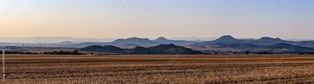 Panorama of Central Bohemian Uplands (Ceske stredohori) during sunset on a golden harvested field, town and factory in the background