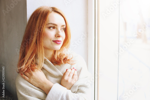 Beautiful redhead young woman with red lipstick on lips sitting by the window of coffee shop, wearing winter autumn season white knitted sweater & big loose scarf on shoulders. Background, close up.