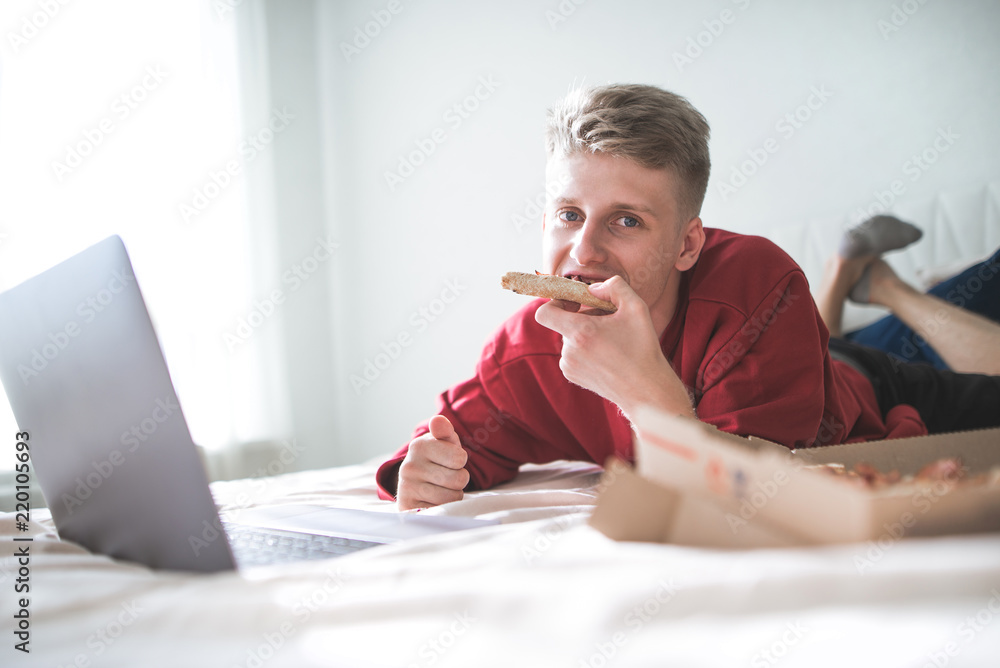 Portrait of an attractive young man lying on the bed with a laptop and a pizza box, eating a piece of pizza and looking at the camera. Teen feeds on pizza at home while watching a movie on a laptop. Portrait of an attractive young man lying on the bed with a laptop and a pizza box, eating a piece of pizza and looking at the camera. Teen feeds on pizza at home while watching a movie on a laptop.