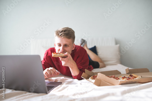 Foto Young happy man is lying on the bed with a box of pizza, uses a laptop and eats a piece of pizza