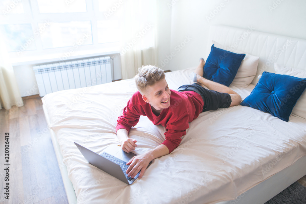 Happy young man lying in bed in a bright cozy room, using a laptop and smiling. Smiling man in a red sweatshirt is working on a laptop in bed and looking at side. Work on the Internet. Happy young man lying in bed in a bright cozy room, using a laptop and smiling. Smiling man in a red sweatshirt is working on a laptop in bed and looking at side. Work on the Internet.