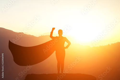 silhouette of a business woman superhero with a cloak standing on top of a mountain in a pose of success in the sunlight