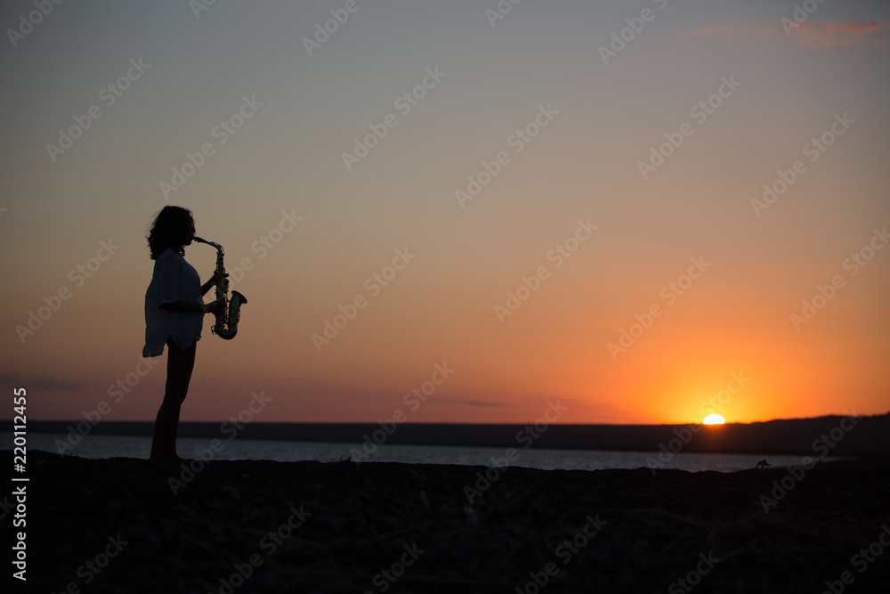 Silhouette of young sexy woman playing saxophone on the beach at sunset