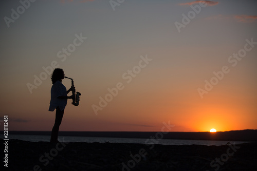 Silhouette of young sexy woman playing saxophone on the beach at sunset