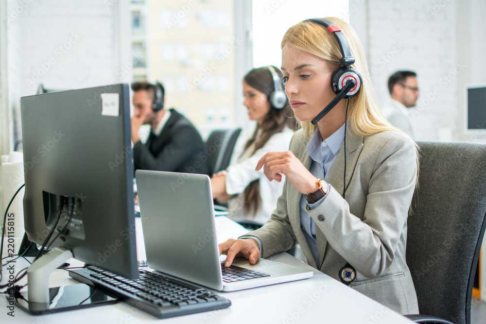 Pensive blonde businesswoman with headset working on laptop in call center Pensive blonde businesswoman with headset working on laptop in call center