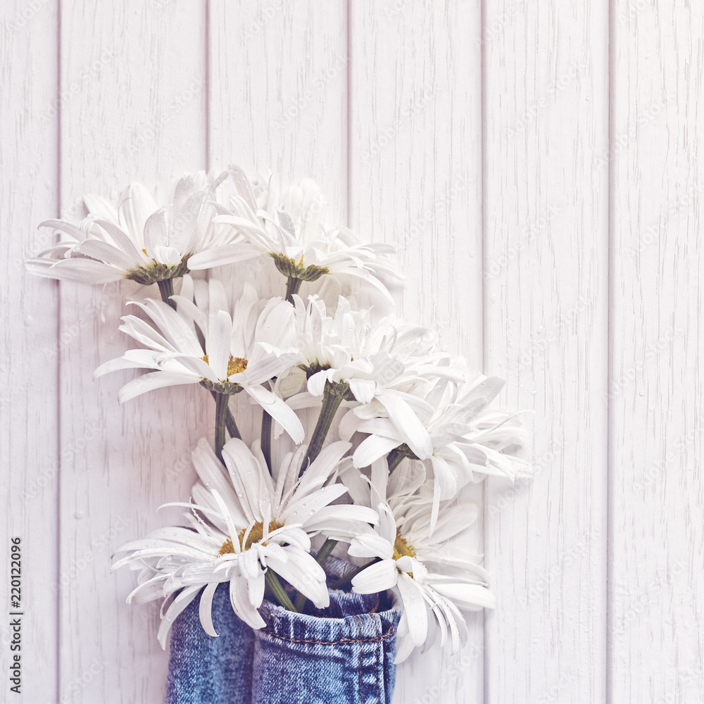 Obraz premium A bouquet of seven white daisies is wrapped in denim fabric. On the petals of flowers there are droplets of water. White wooden background.