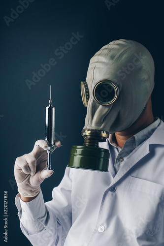Creepy medical experiment concept, a scary doctor in gas mask with a big stainless steel syringe on dark background
