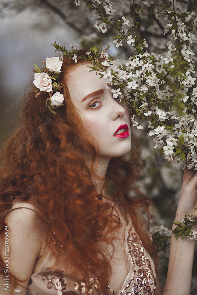 A gentle woman with long red hair in a blooming spring garden. Red sensual girl with pale skin and blue eyes with bright unusual appearance hides behind flowers. A gentle woman with long red hair in a blooming spring garden. Red sensual girl with pale skin and blue eyes with bright unusual appearance hides behind flowers.