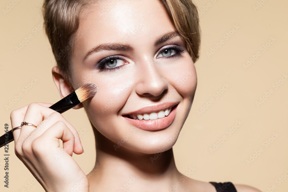 Portrait of beautiful girl with alluring eyes. Sensual female holding in hand cosmetic tool for applying tonal basic. Smiling blonde looking at camera with happiness. Isolated on beige background Portrait of beautiful girl with alluring eyes. Sensual female holding in hand cosmetic tool for applying tonal basic. Smiling blonde looking at camera with happiness. Isolated on beige background