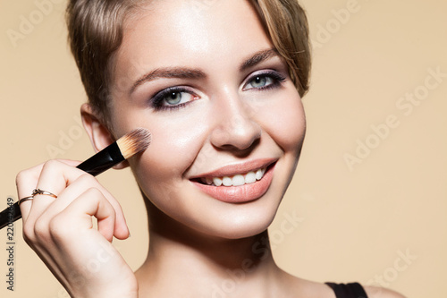 Portrait of beautiful girl with alluring eyes. Sensual female holding in hand cosmetic tool for applying tonal basic. Smiling blonde looking at camera with happiness. Isolated on beige background