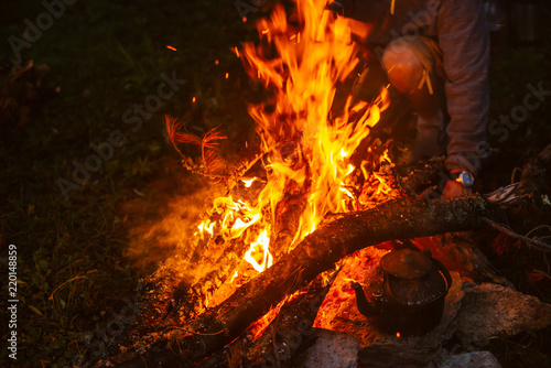 Hot tea in kettle on bonfire. Man bask. Hand throws up brushes in fire. Tea drinking in open air. Active outdoor recreation. Camping in dusk. Atmospheric warm in twilight. Active rest.