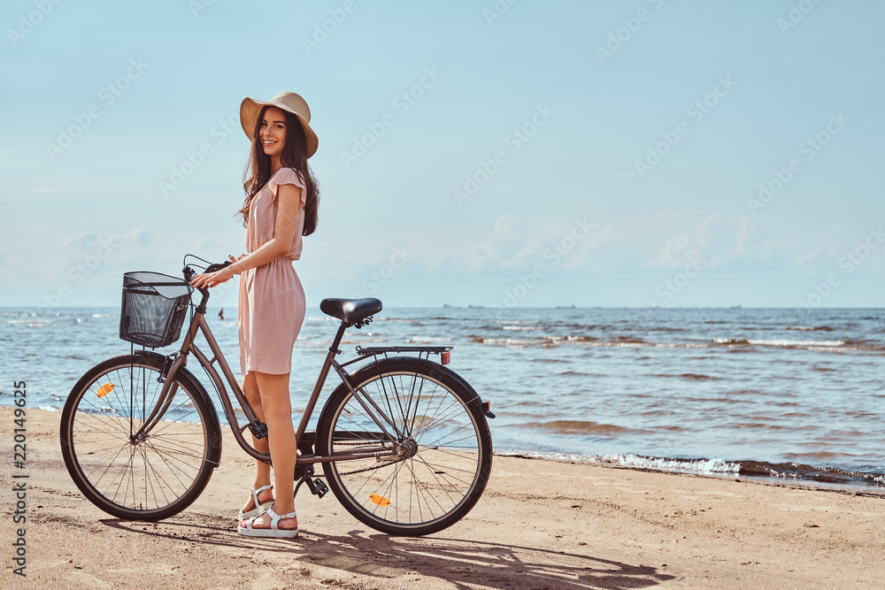 Fototapeta premium Beautiful brunette girl dressed in dress and hat posing with a bicycle on the beach on a sunny day.