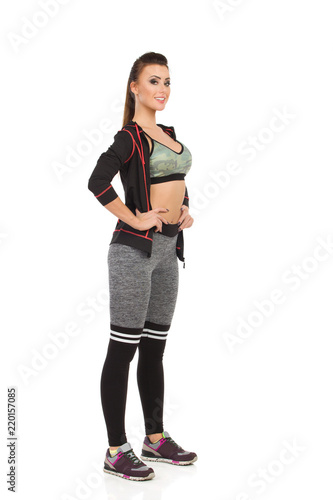 Beautiful Young Woman In Sports Clothes Is Standing With Hand On Hip And Looking At Camera. Front Side View.