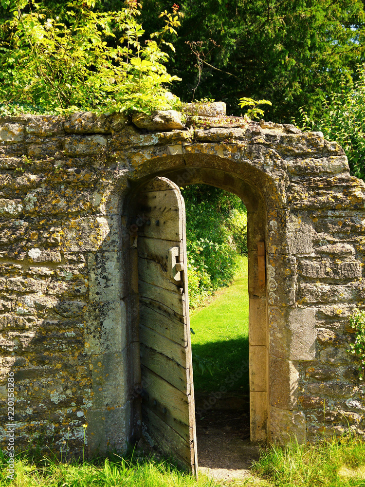 An open wooden door in an ancient ruined wall, leads to a secret garden in the Cotswolds England.