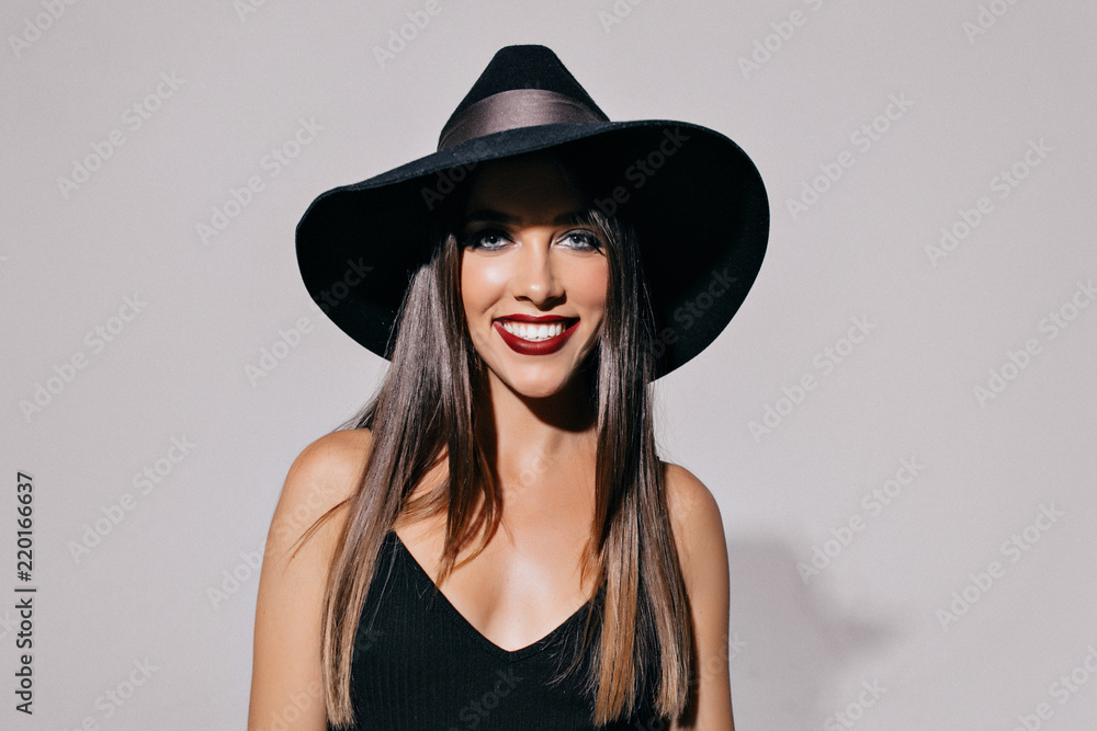 Studio shot of attractive pretty young woman with smoky eyes and black lips wearing hat and black dress posing front of the wall. Halloween, masquerade, party, celebration Studio shot of attractive pretty young woman with smoky eyes and black lips wearing hat and black dress posing front of the wall. Halloween, masquerade, party, celebration