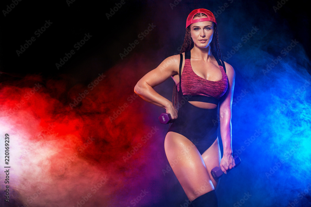 Obraz premium Attractive young fitness woman athlete and bodybuilder holding dumbell on black background with color smoke. Copy space for fitness nutrition ads.