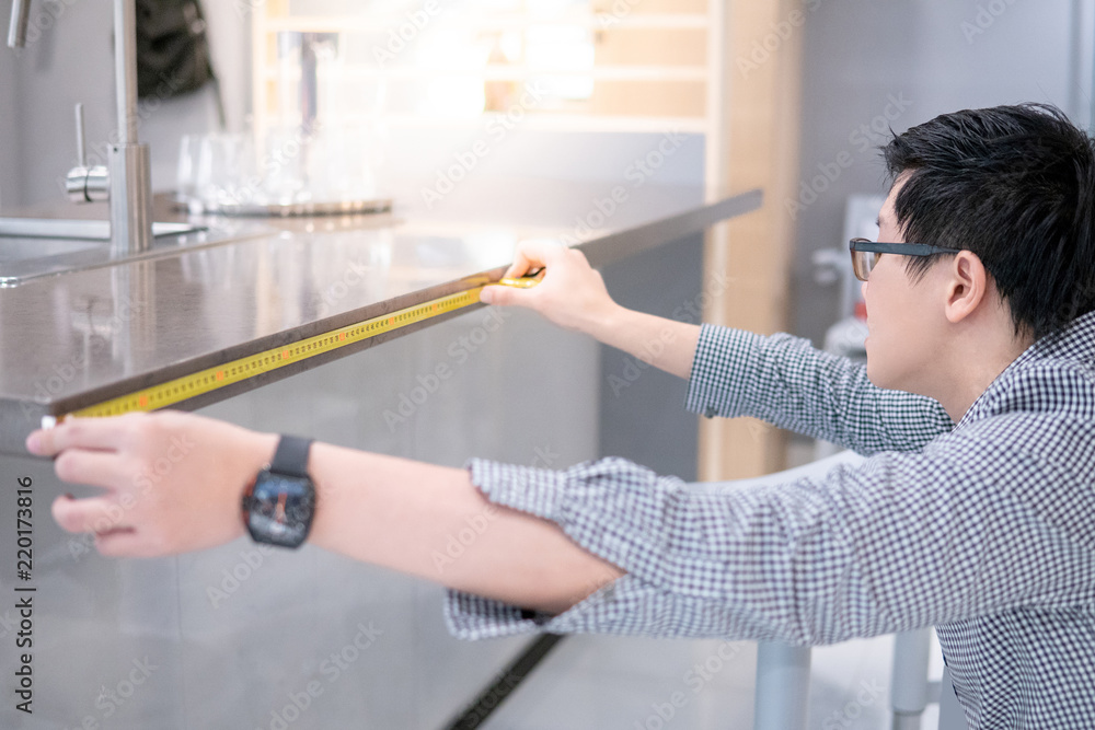 Young Asian man using tape measure for measuring granite countertops on modern kitchen counter in showroom. Shopping furniture for home improvement. Interior design concept Young Asian man using tape measure for measuring granite countertops on modern kitchen counter in showroom. Shopping furniture for home improvement. Interior design concept