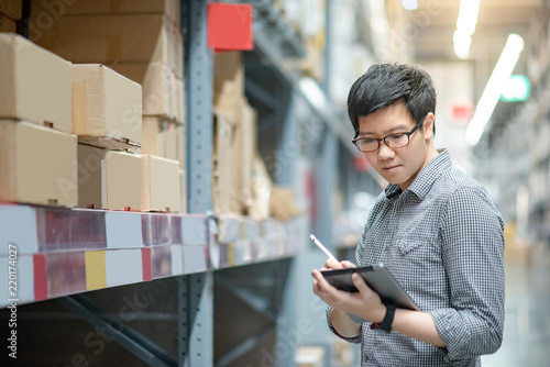 Young Asian man worker doing stocktaking of product in cardboard box on shelves in warehouse by using digital tablet and pen. Physical inventory count concept photo