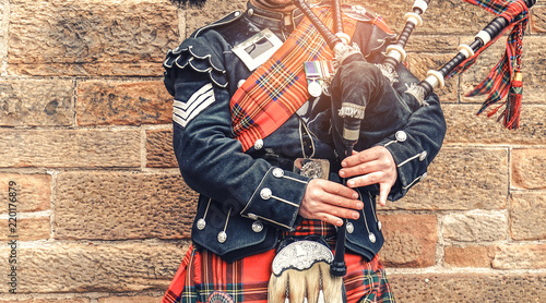 Fotografia EDINBURGH, SCOTLAND, 24 March 2018 , Scottish bagpiper dressed in traditional red and black tartan dress stand before stone wall