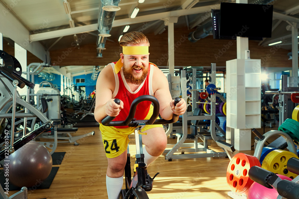 A funny fat man with a beard doing exercises on an exercise bike in a  sports hall. Stock Photo | Adobe Stock