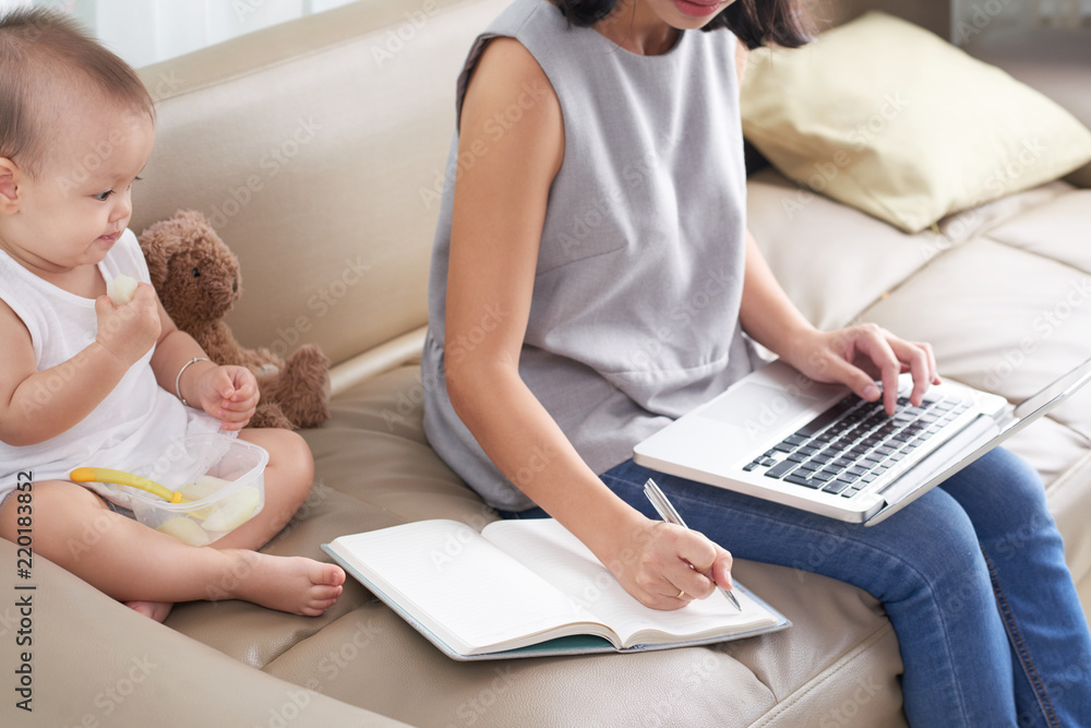 Obraz premium Young mother working on laptop and taking notes when her baby eating